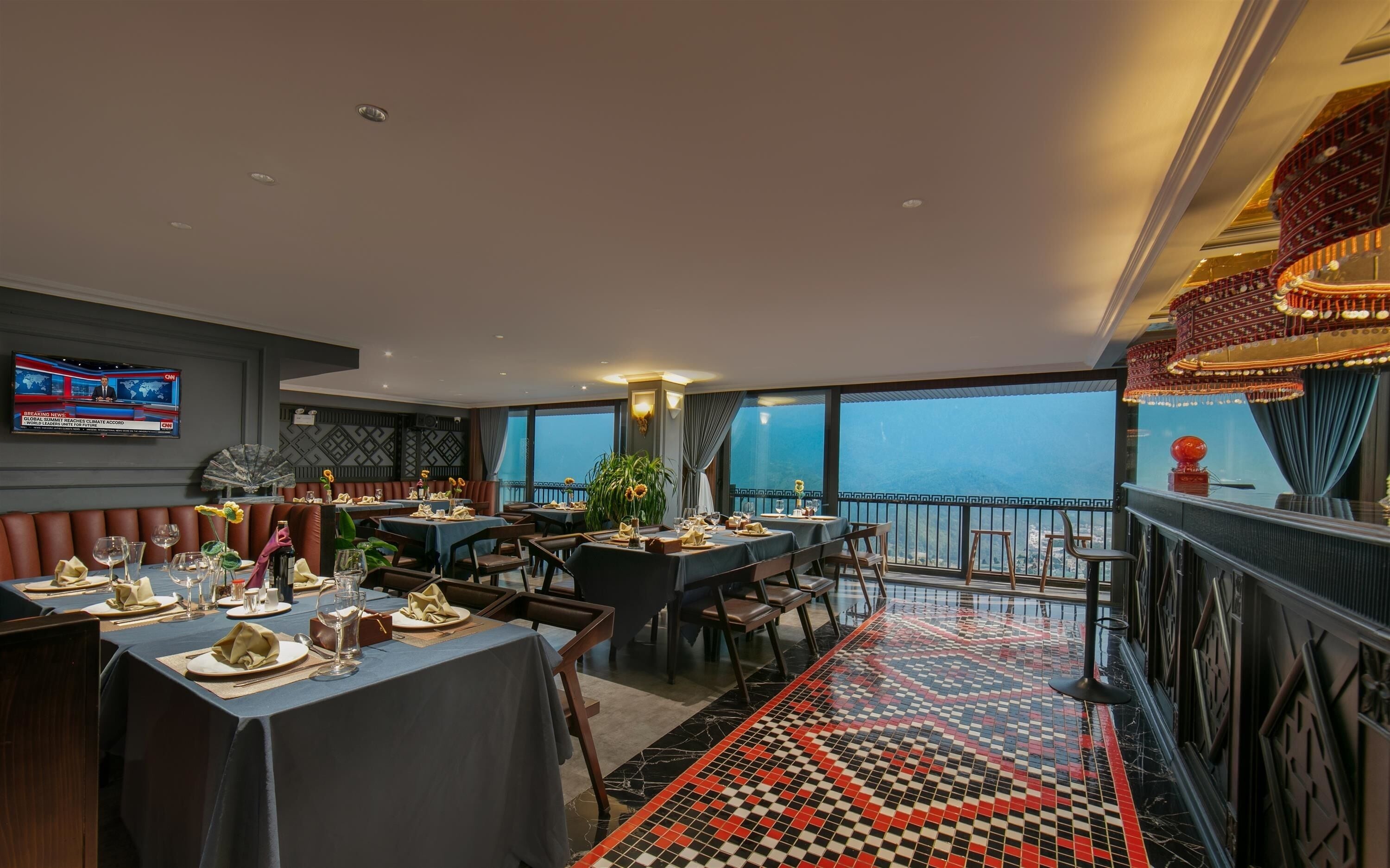 A restaurant with tables set for dining, a TV screen displaying news, and a balcony with a view.