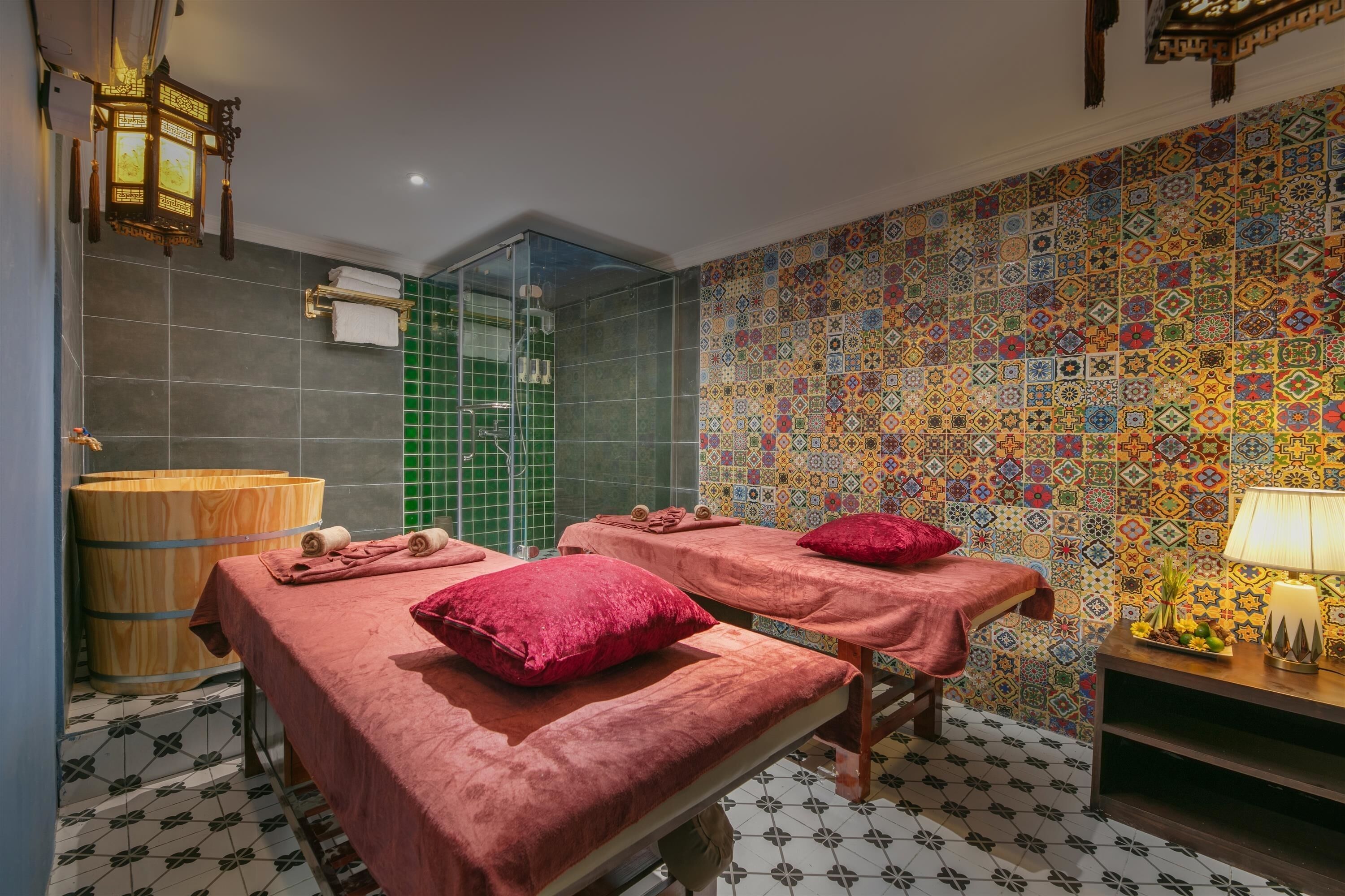 A room with two massage tables, a wooden barrel, a green-tiled shower area, and a colorful patterned wall.