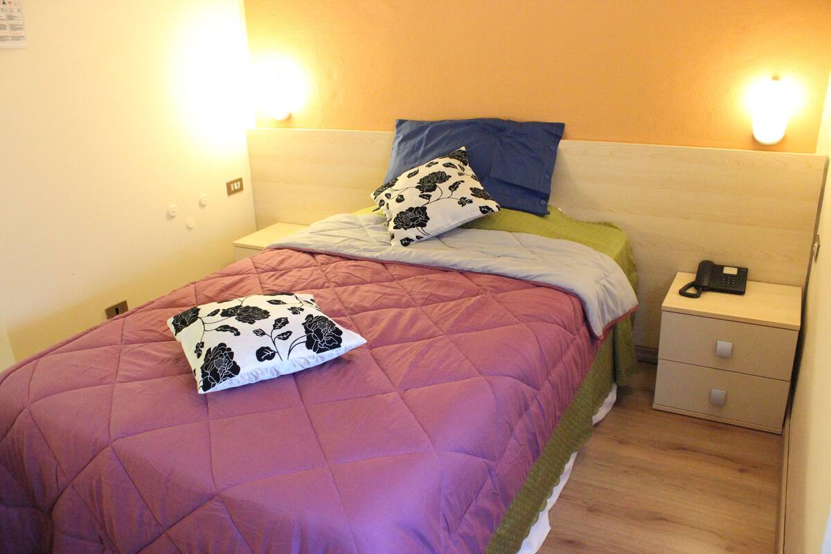 A neatly made bed with a purple quilt, a blue pillow, and a white pillow with black floral patterns. There is a nightstand with a phone and a lamp beside the bed.