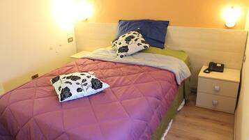 A neatly made bed with a purple quilt, a blue pillow, and a white pillow with black floral patterns. There is a nightstand with a phone and a lamp beside the bed.