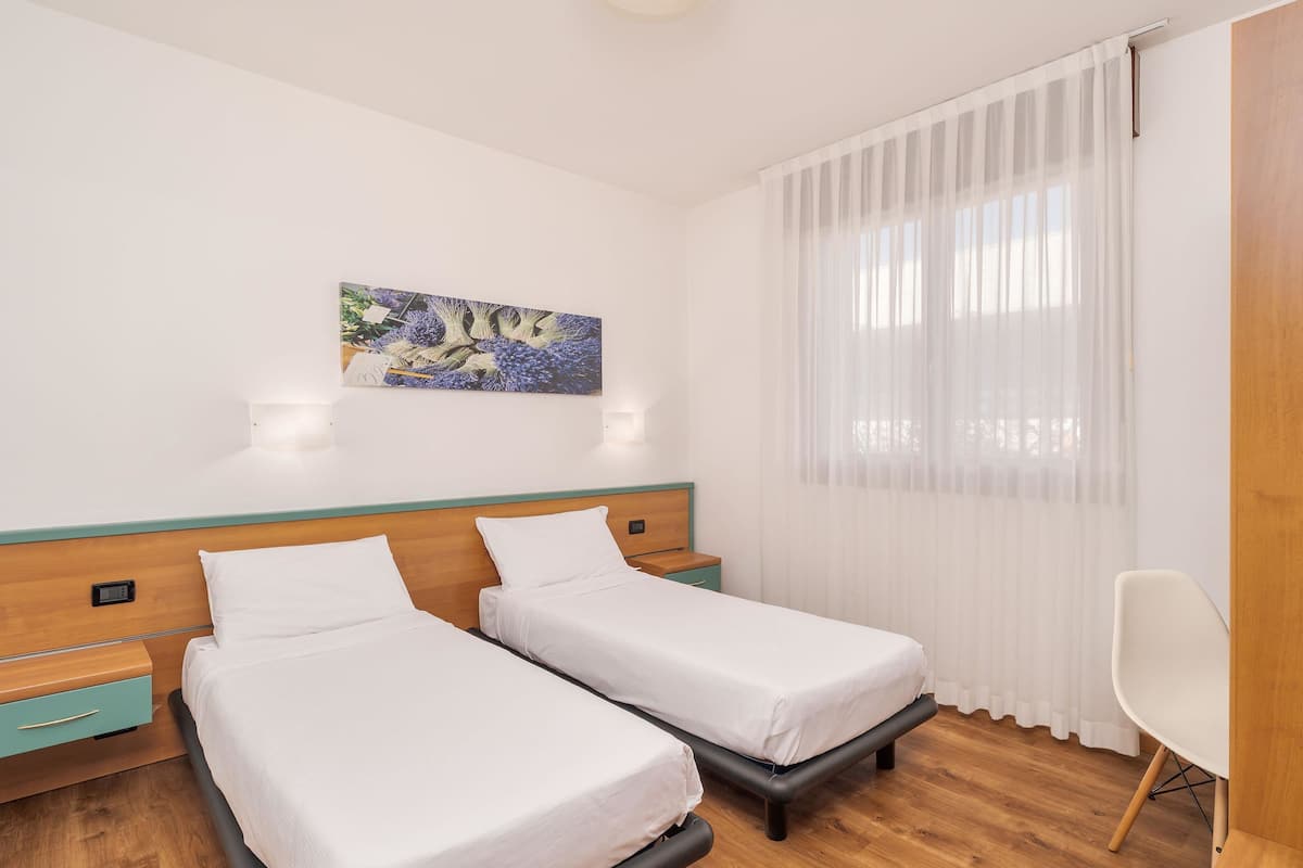 A hotel room with two single beds, a wooden headboard, a small bedside table with a lamp, a chair, and a painting on the wall.