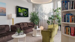A modern living room with a brown sofa, a green armchair, a white coffee table, a bookshelf filled with books, and a large window with sheer curtains. - B&B Hotel Eurorest Conegliano (Conegliano)