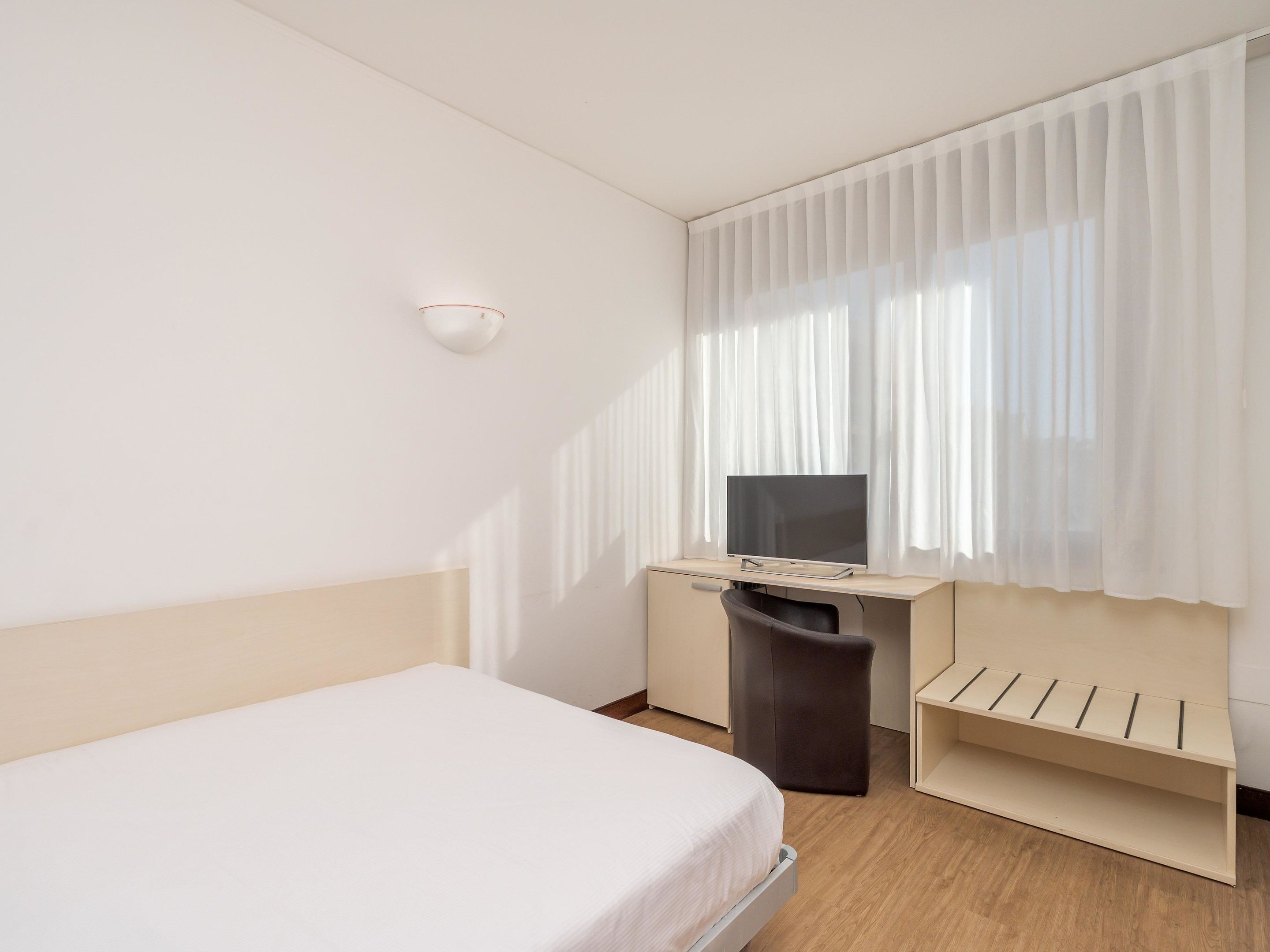 Une chambre d’hôtel avec un lit, un bureau avec une télévision, une poubelle et une fenêtre avec des rideaux.