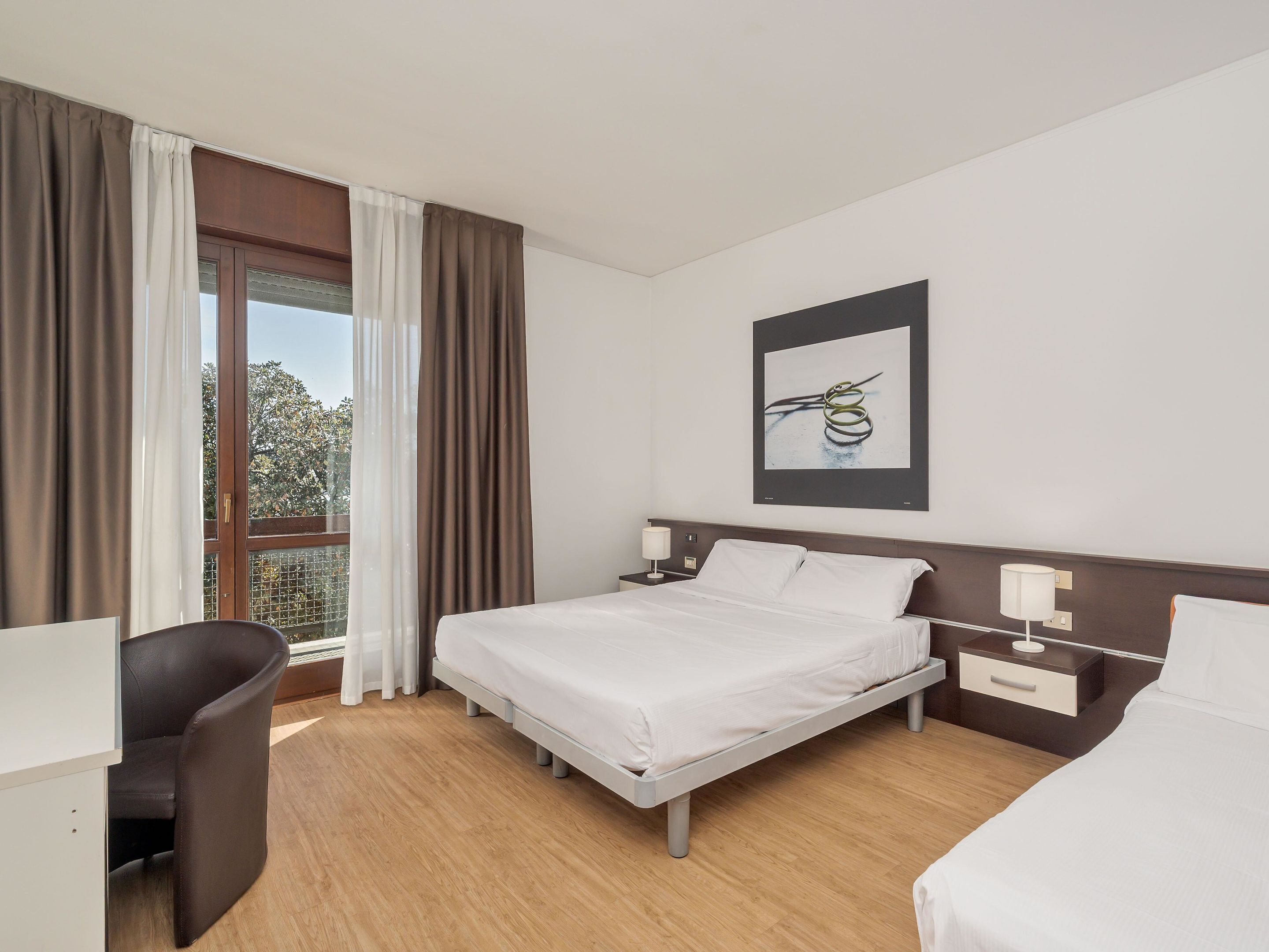 Ein Hotelzimmer mit Bett, Schreibtisch und Stuhl. Es gibt ein Fenster mit Vorhängen, ein gerahmtes Bild an der Wand und einen Nachttisch mit Lampe.