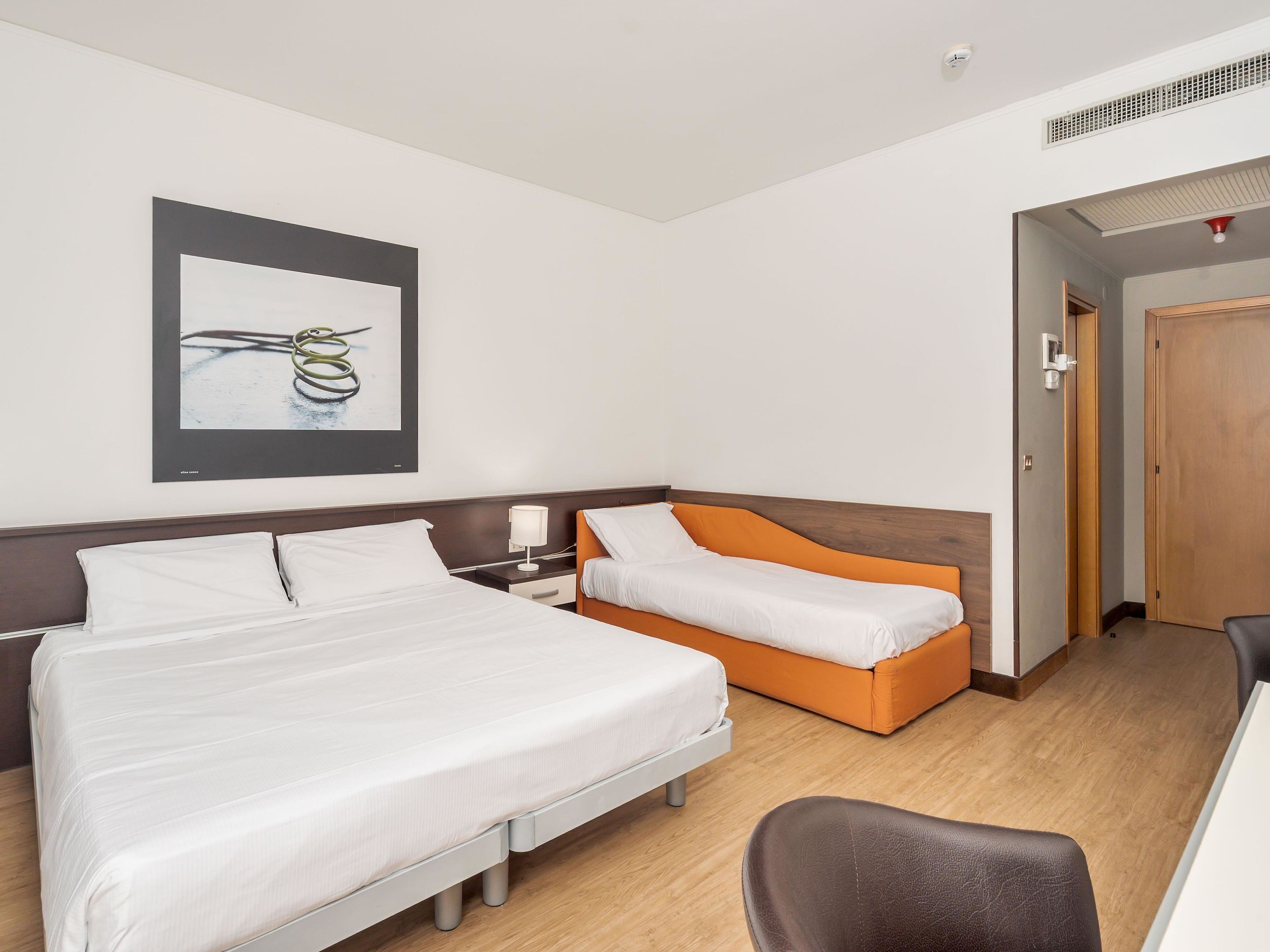 A hotel room with two beds, one with white bedding and the other with orange bedding, a bedside table with a lamp, a wooden headboard, a framed picture on the wall, and a wooden door.