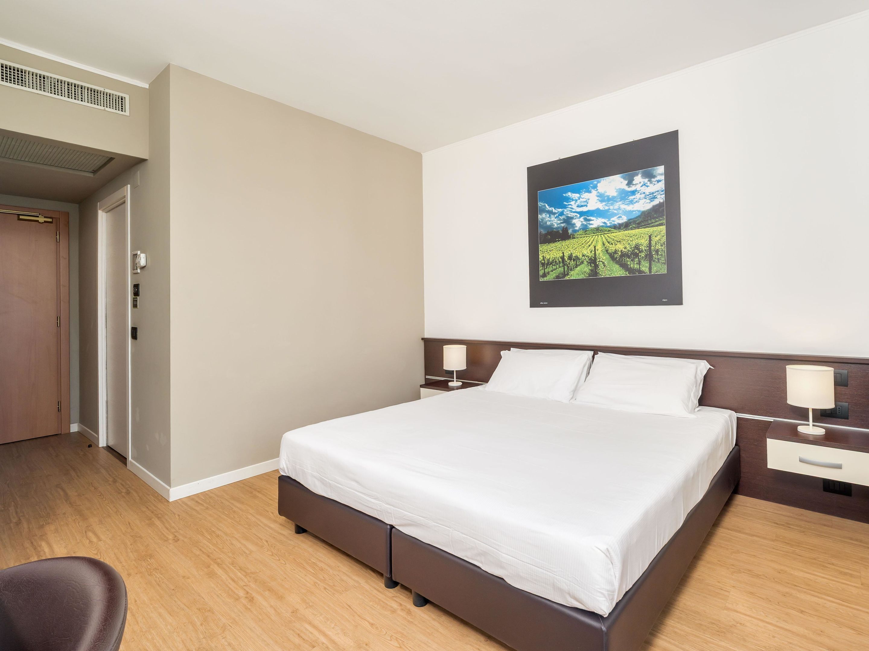 A hotel room with a large bed, a nightstand, a wall-mounted picture, and a door to the left.