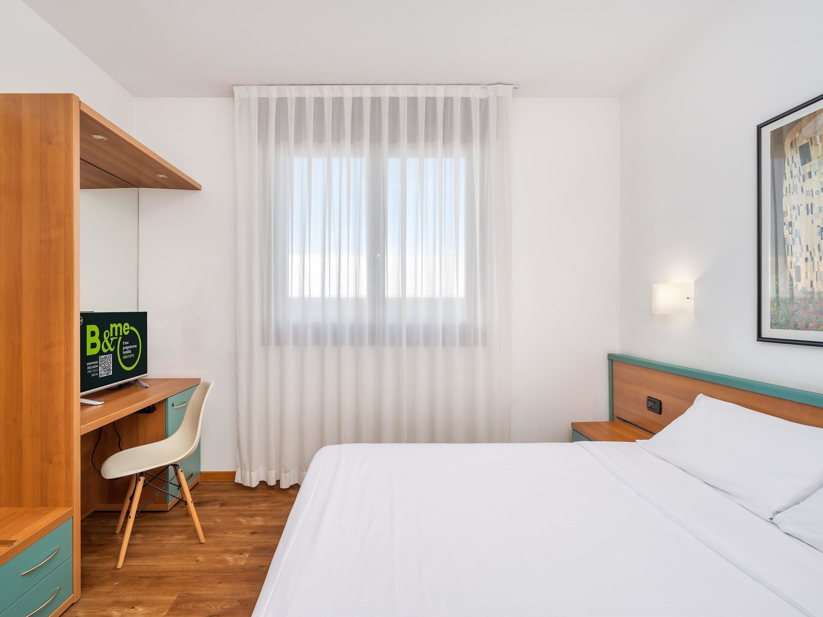 Hotelkamer met een bed, een bureau, een stoel, een kledingkast en een raam met dunne gordijnen.