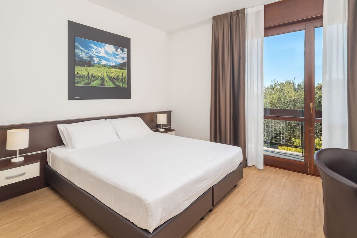 A hotel room with a large bed, a nightstand, a lamp, a picture on the wall, and a balcony with a view of greenery.
