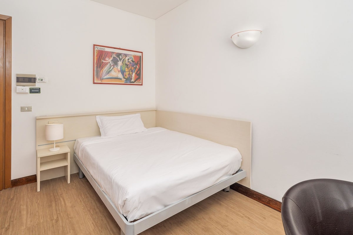 A hotel room with a bed, a bedside table with a lamp, a wall-mounted light, and a painting.