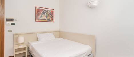 A hotel room with a bed, a bedside table with a lamp, a wall-mounted light, and a painting.