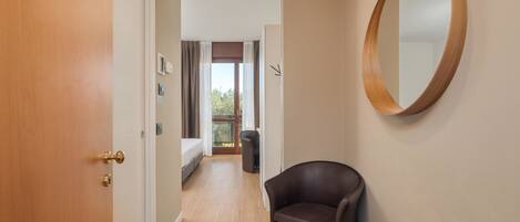 A hotel room with a wooden door, a small round table, a leather chair, a bed, and a balcony with curtains.