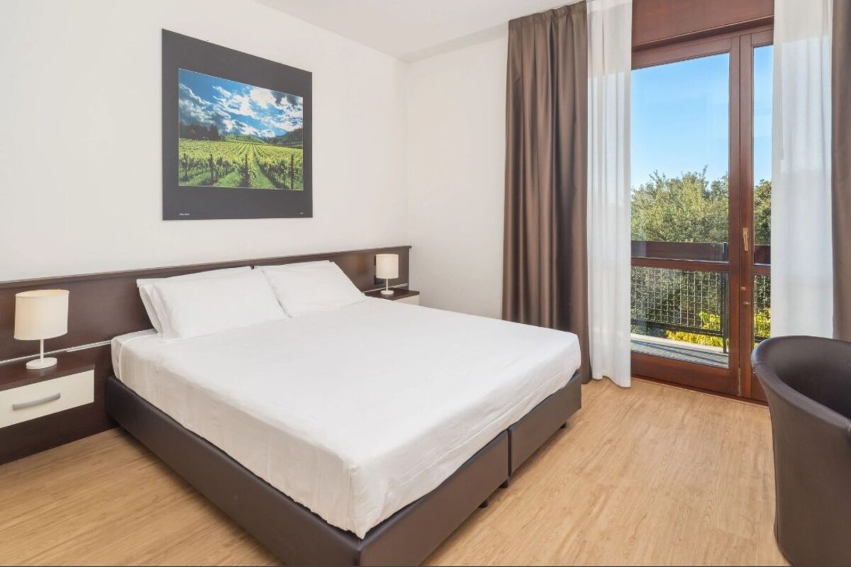 Una habitación de hotel con cama, mesita de noche, lámpara, un cuadro de un viñedo y un balcón con cortinas.