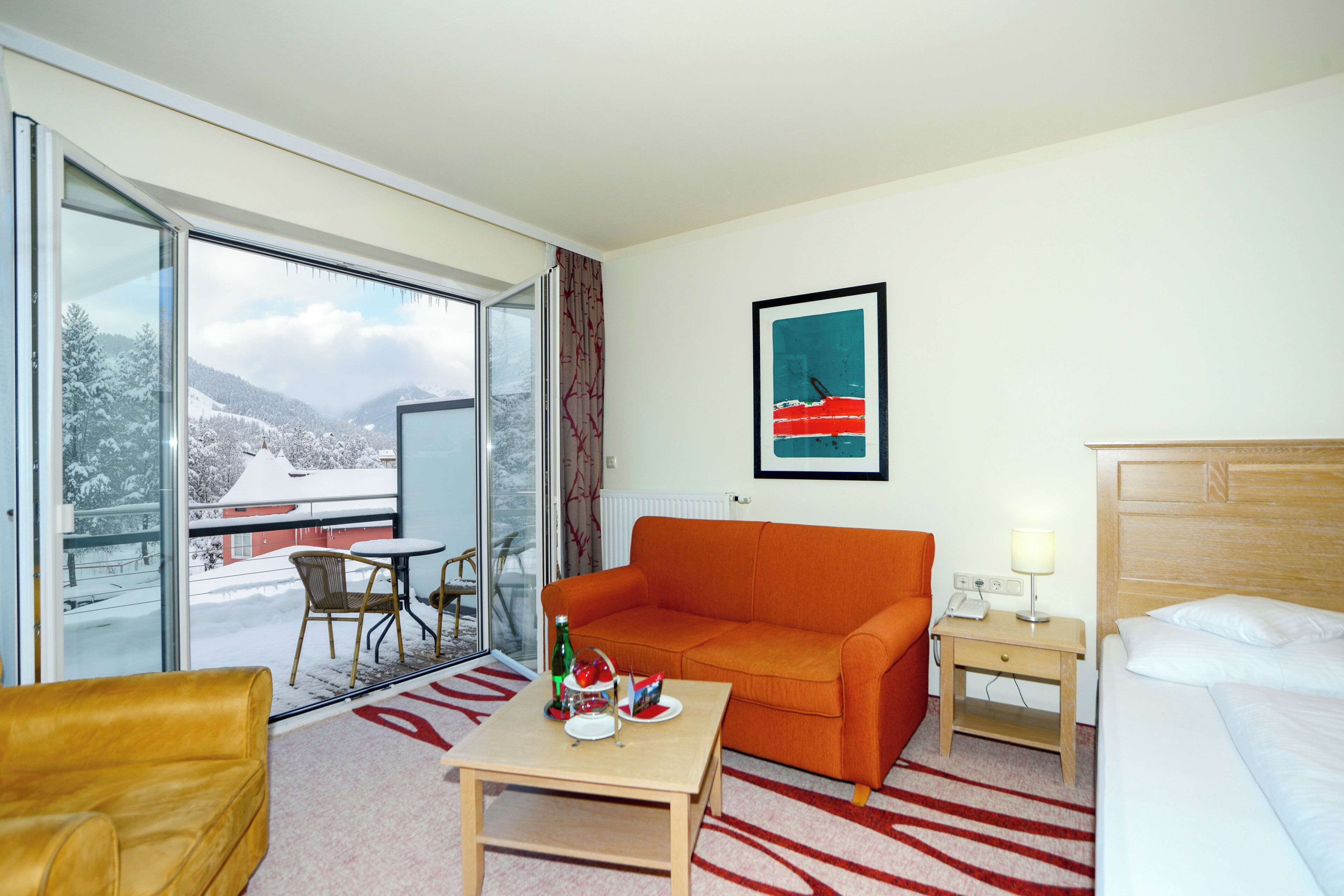 A hotel room with a sofa, a small table with a bottle and glasses, a bed, a nightstand with a lamp, a framed picture on the wall, and a view of snowy mountains through the glass door.