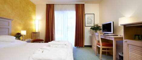 Quarto de hotel com uma cama, uma escrivaninha com computador, uma cadeira e uma janela com cortinas.
