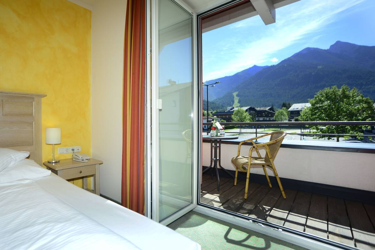 A hotel room with a bed, a nightstand with a lamp, a phone, and a table with a chair. The room has a view of mountains and a balcony with a table and chair.