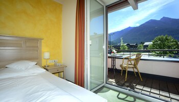 Habitación de hotel con cama, mesita de noche con lámpara, teléfono y una mesa con silla. La habitación tiene vistas a montañas y un balcón con mesa y sillón.