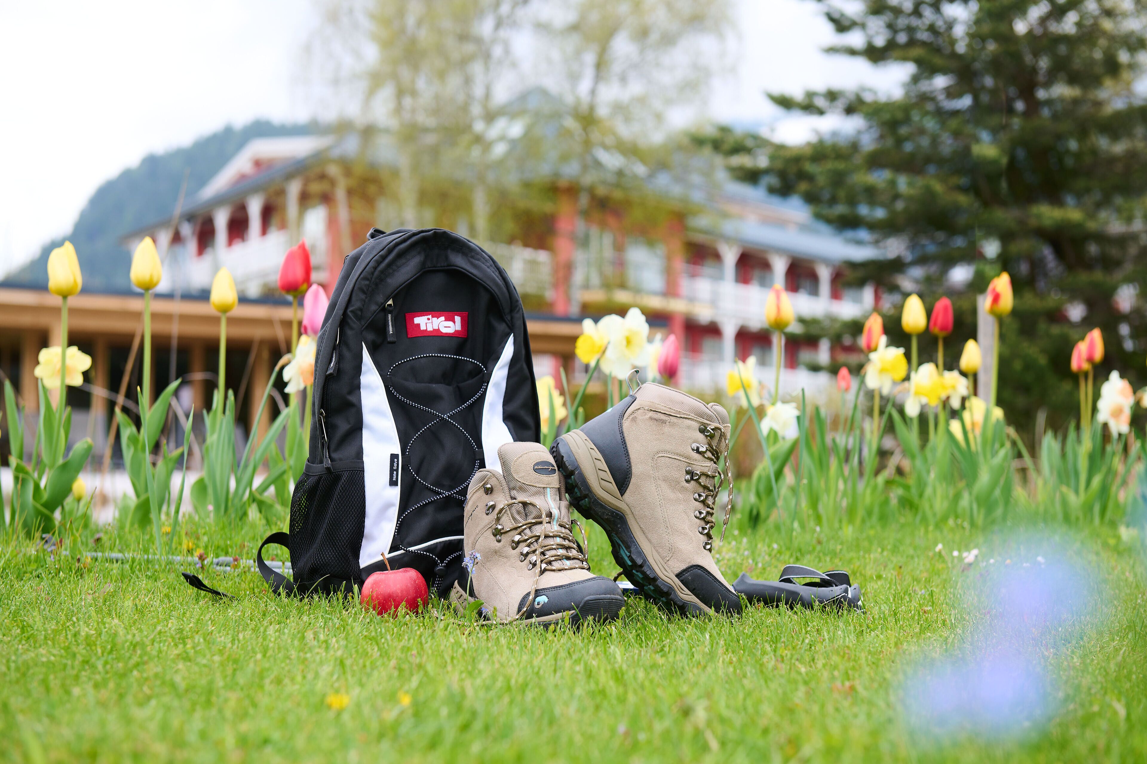 A pair of hiking boots, a black backpack, and a red ball on a grassy field with tulips and a building in the background.