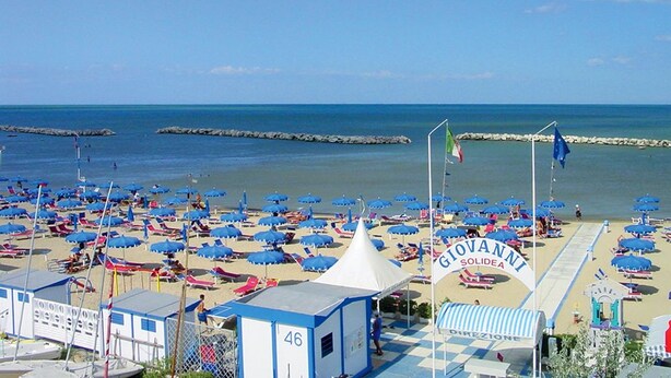 Une plage parsemée de nombreux parasols bleus et de chaises longues, une tente blanche et une pancarte sur laquelle on peut lire « Giovanni Solidea ».