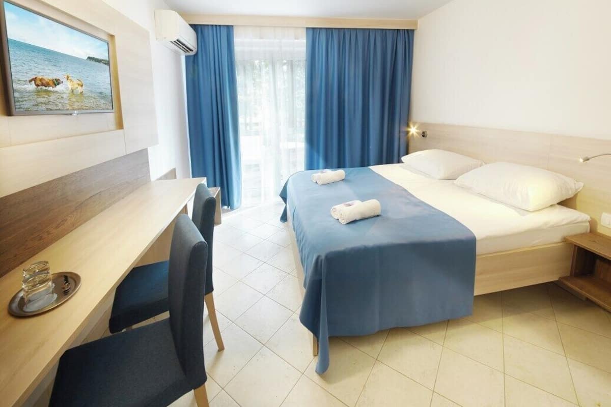 A hotel room with a bed, a desk, and a chair. There is a TV mounted on the wall, and a window with curtains.