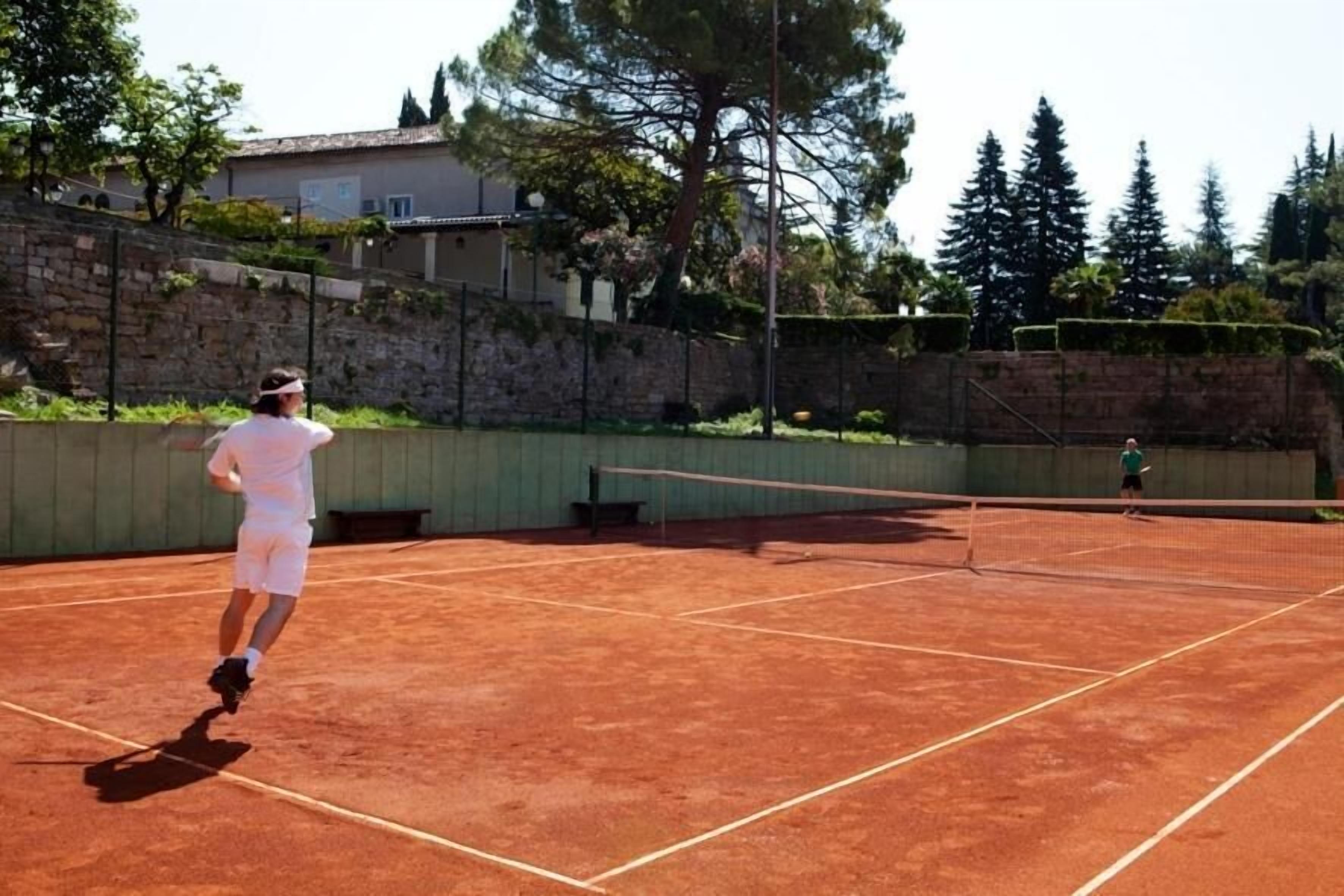 A tennis court with a net in the center, surrounded by a stone wall and trees.