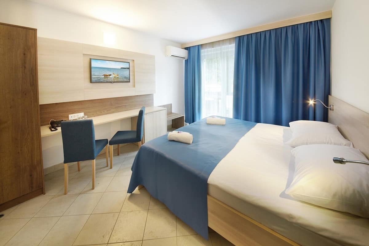 A hotel room with a bed, a desk, a chair, a painting on the wall, and a window with curtains.