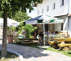 Área externa com mesas e cadeiras, um alpendre coberto e um edifício ao fundo.