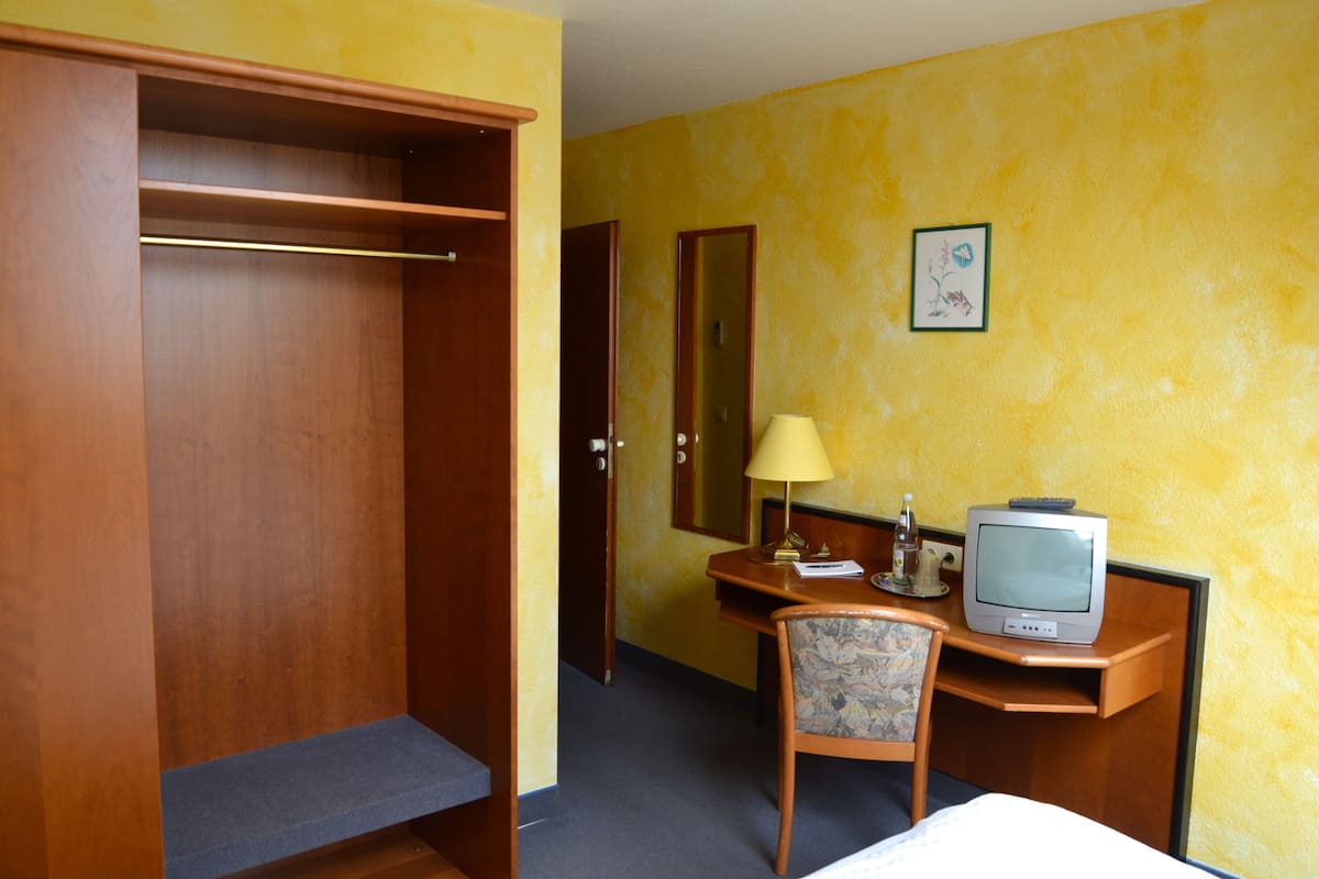 A hotel room with a wooden wardrobe, a desk with a television, a chair, and a bed.