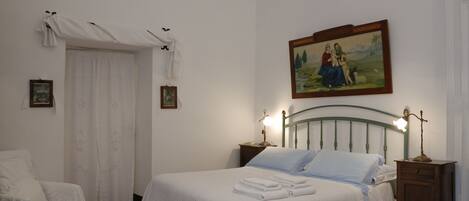 Een netjes opgemaakt slaapkamer met een bed, nachtkastje, stoel en een schilderij aan de muur.