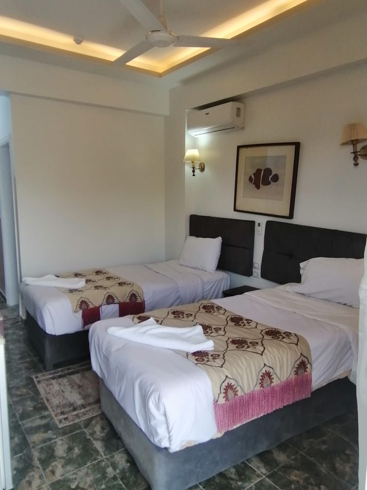 Habitación de hotel con dos camas, un ventilador de techo, un equipo de aire acondicionado, un cuadro enmarcado y una lámpara fijada a la pared.
