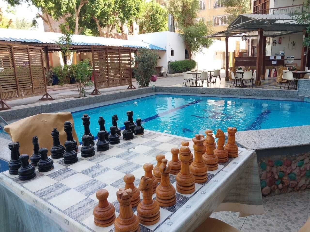 A poolside chessboard with black and brown pieces, surrounded by a pool, outdoor seating, and a building in the background.