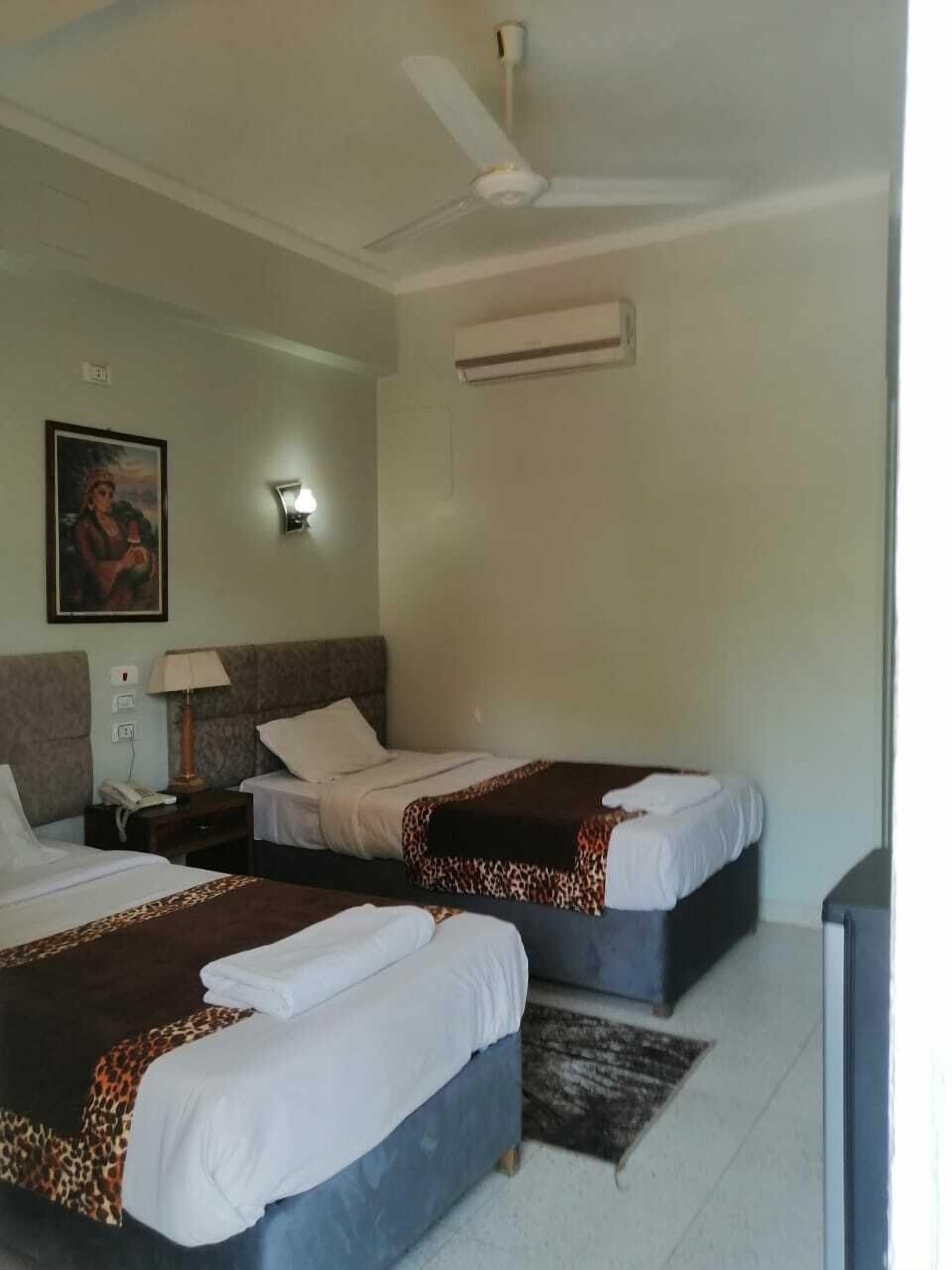 A hotel room with two beds, a ceiling fan, an air conditioning unit, a painting on the wall, a bedside lamp, and a television.