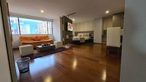 A modern living room with a sofa, ottoman, and a coffee table. There is a TV mounted on the wall and a bed in the background. - Hotel Confort 80 Zona Rosa (Bogotá)