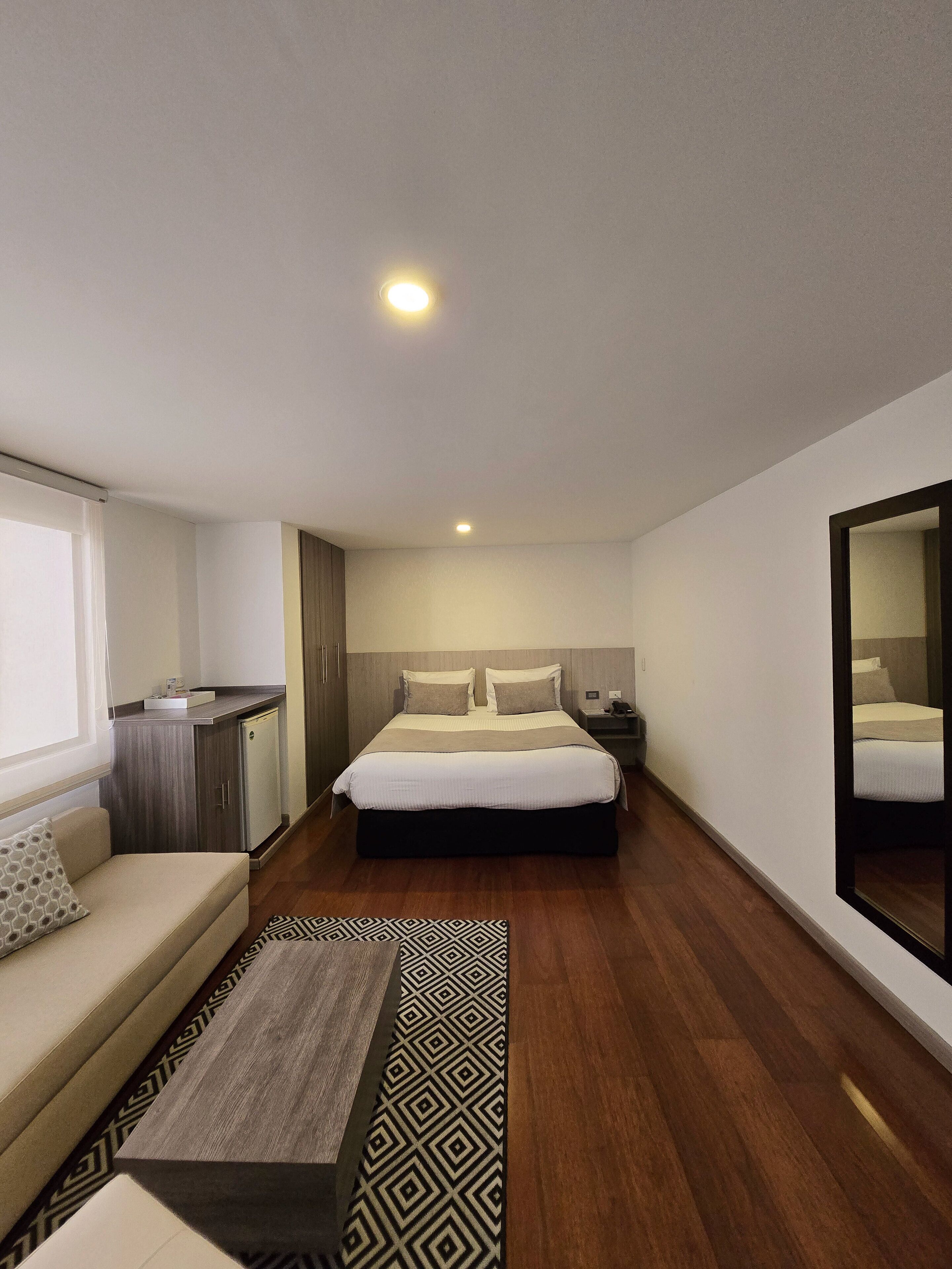 A modern hotel room with a large bed, a sofa, a small kitchenette, and a large mirror.