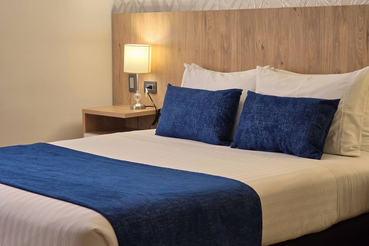 Una habitación de hotel con cabecera de madera, una mesita de noche con lámpara y dos almohadas.