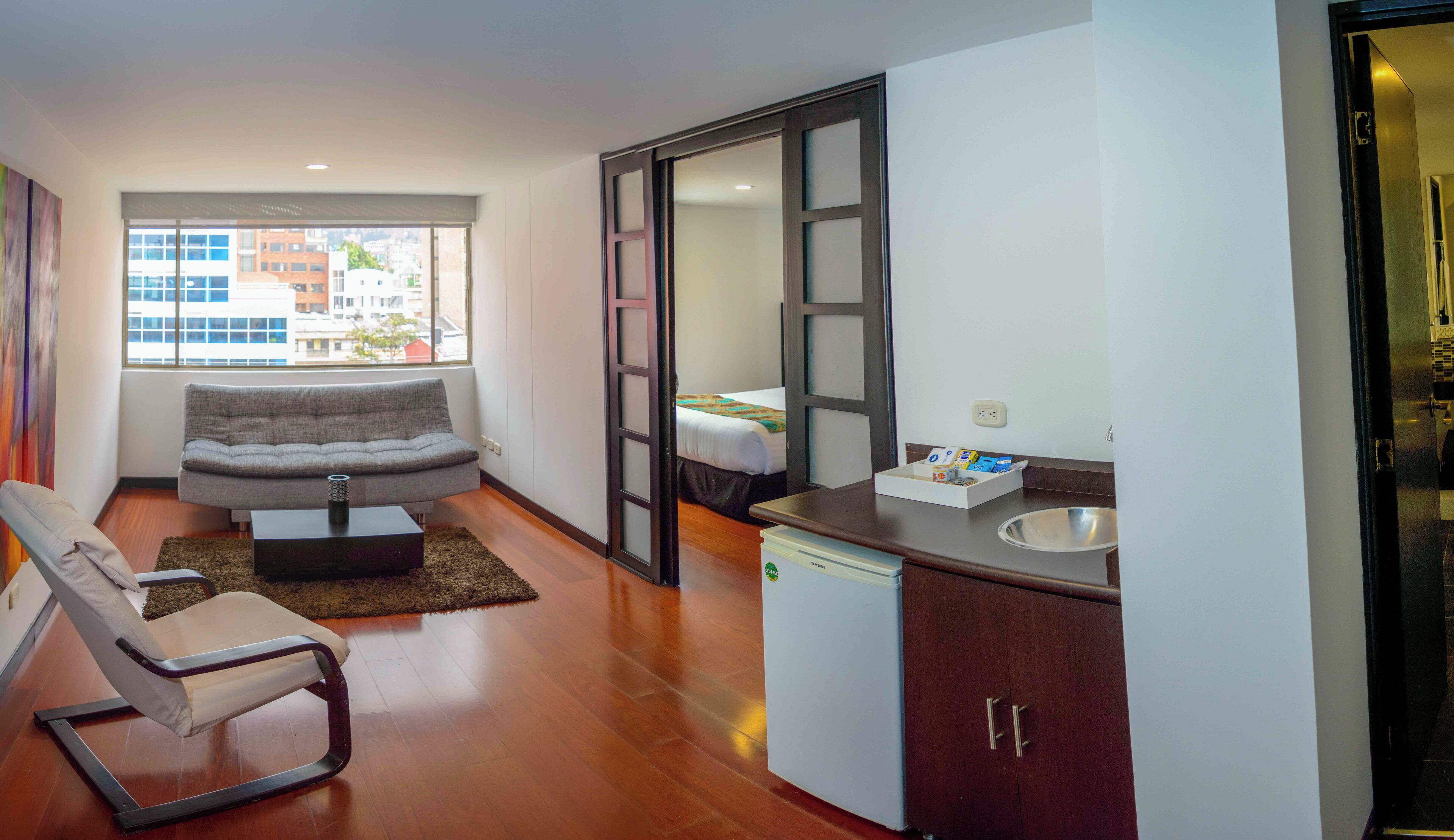 A modern living room with a grey sofa, a small table, and a chair. There is a kitchen area with a sink and cabinets.