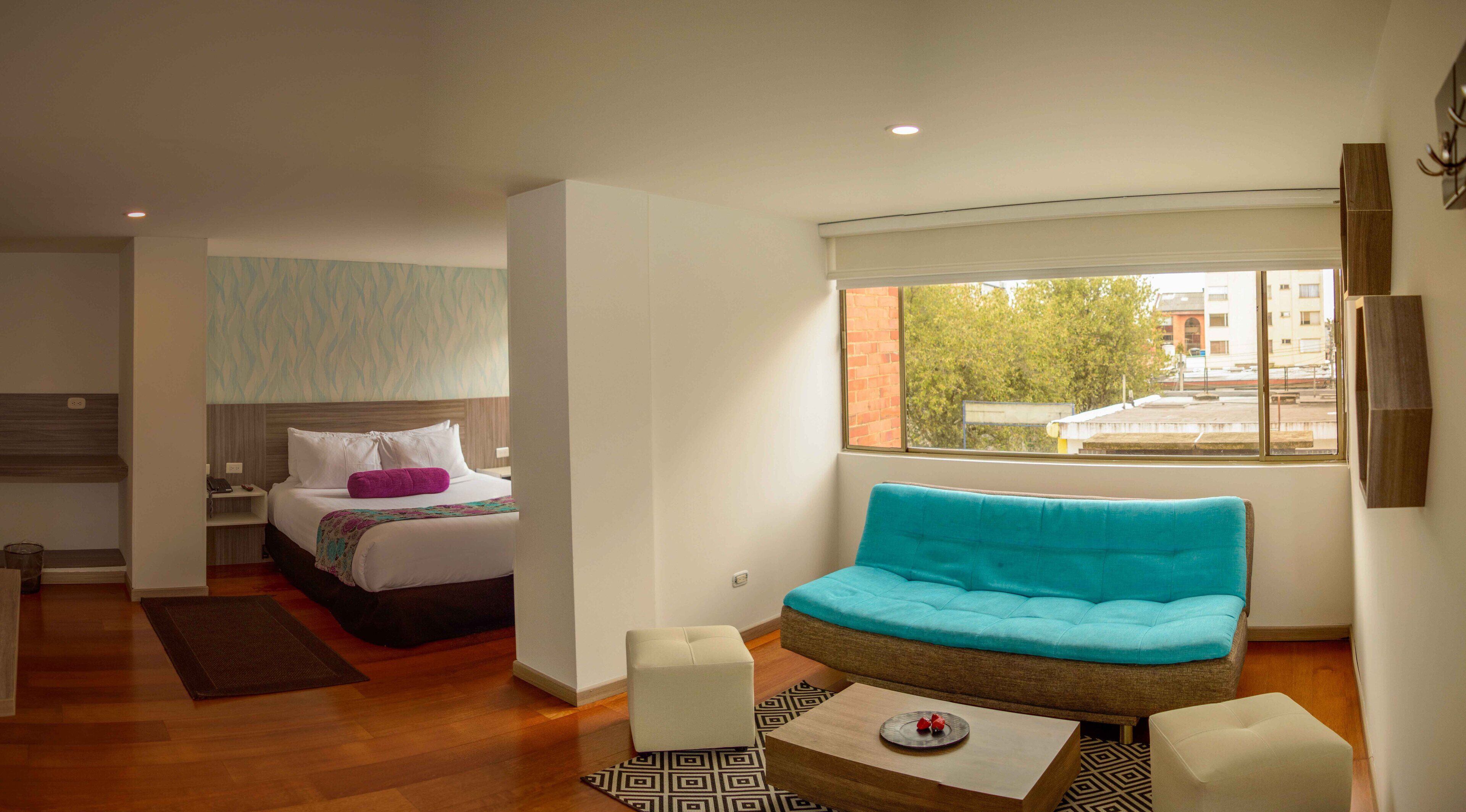A modern hotel room with a bed, a sofa, a small table, and a window with a view of buildings and trees.