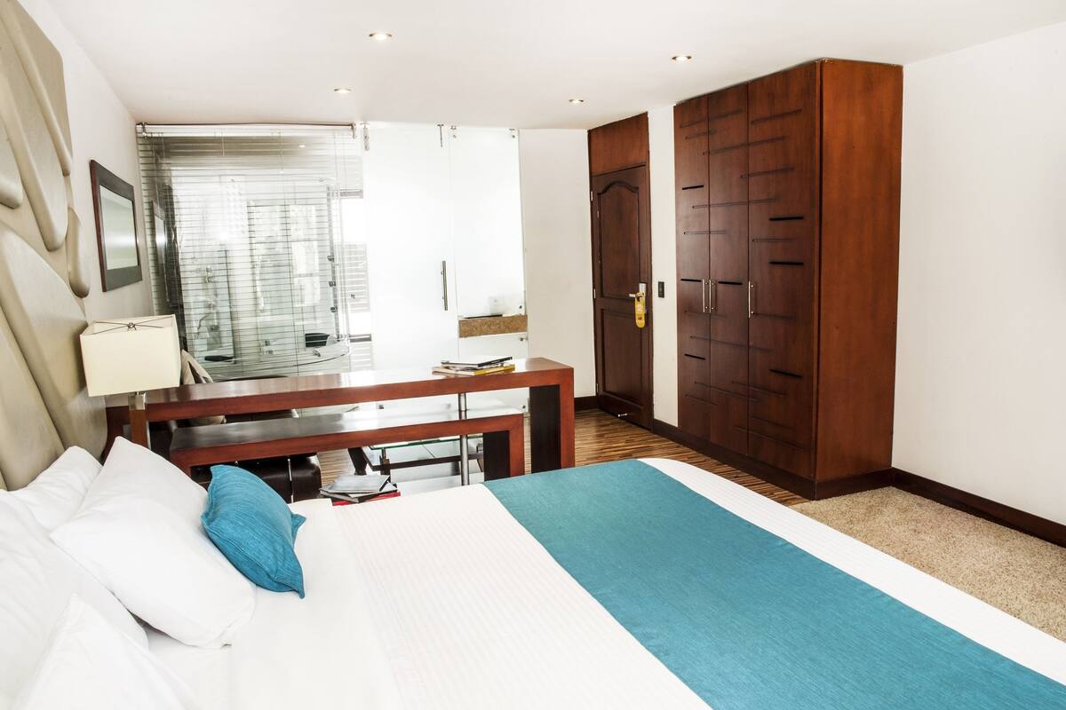 Habitación de hotel con cama, escritorio, armario y baño visible a través de una puerta de cristal.