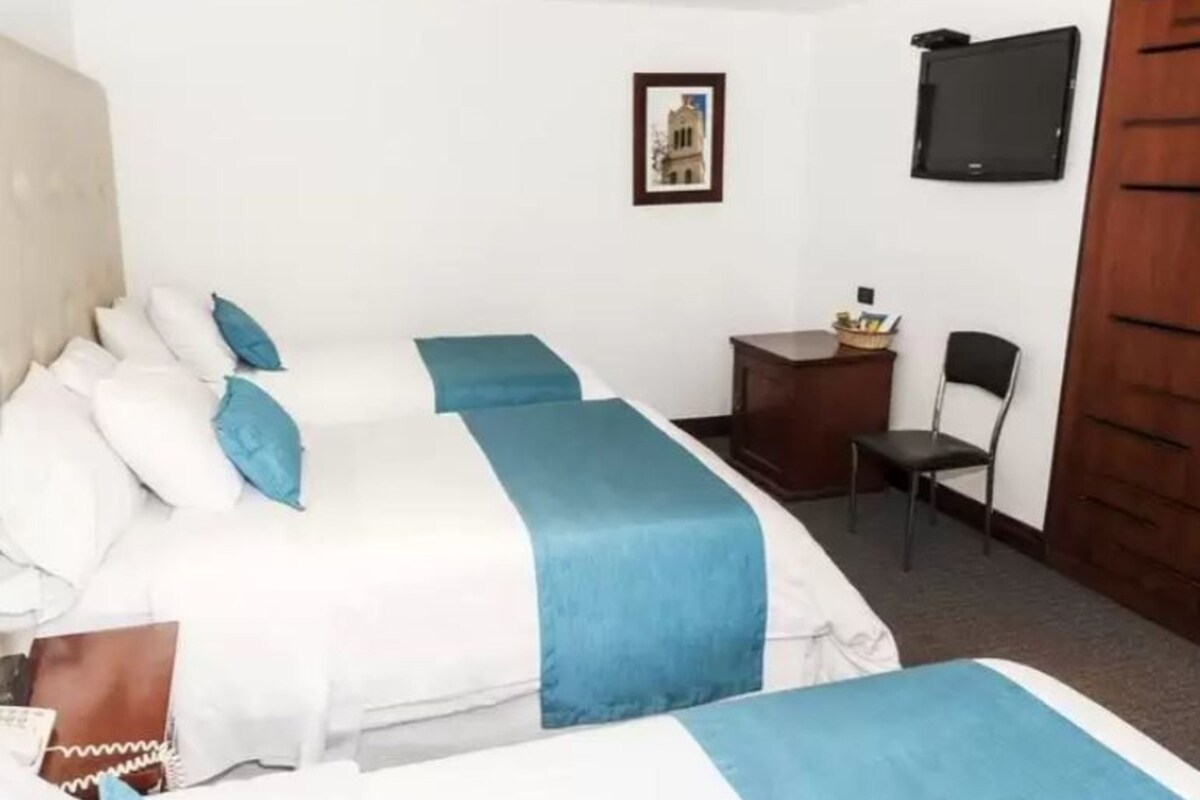 Une chambre d’hôtel avec deux lits, chacun agrémenté de draps blancs et bleus, un petit bureau avec une chaise, un téléviseur fixé au mur et un tableau encadré.