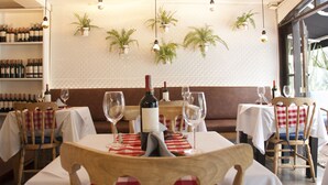 Un interior de restaurante con mesas dispuestas para comer, botellas de vino en estantes y plantas colgantes en la pared.