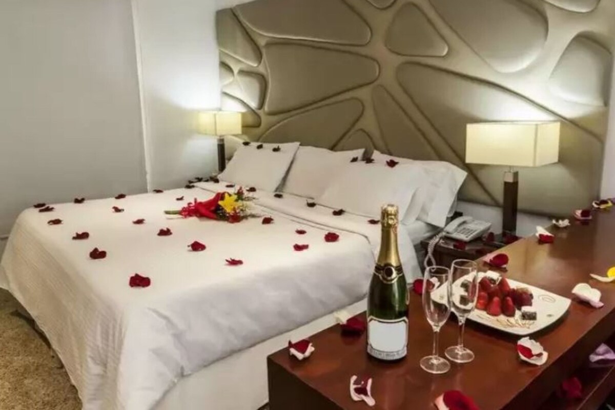 Une chambre d’hôtel avec un lit parsemé de pétales de rose, une bouteille de champagne et une assiette de fruits frais.