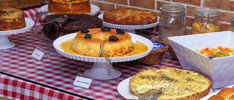 A buffet table with various cakes and pastries, a sign indicating 'Pão de Leite de Laleão', and a bowl of vegetables.
