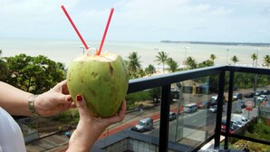 Una mano sostiene un coco verde con pajitas, con vista a una playa con palmeras y una carretera con coches.