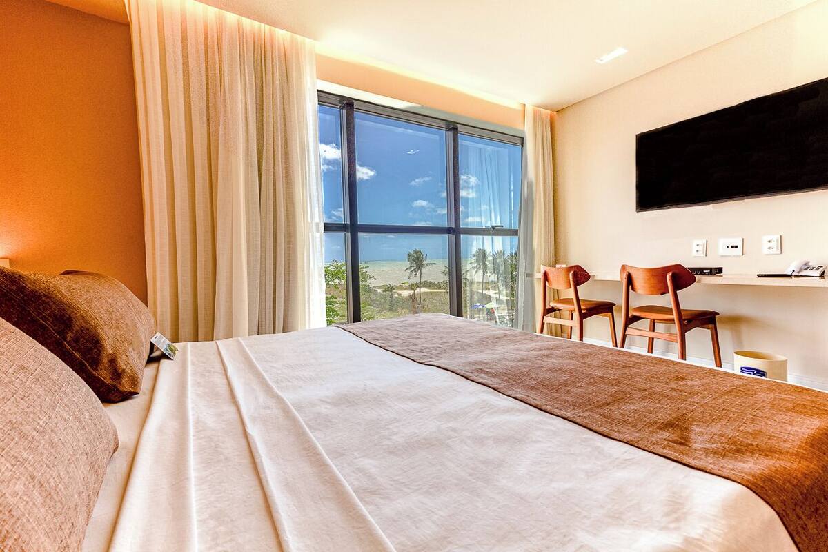 A hotel room with a large bed, a TV mounted on the wall, and a view of the beach through the window.