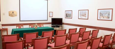 A conference room with a projector screen, a table with a green tablecloth, and several chairs. There are framed pictures on the wall and a sign that reads "HOTEL Il Nachiero".