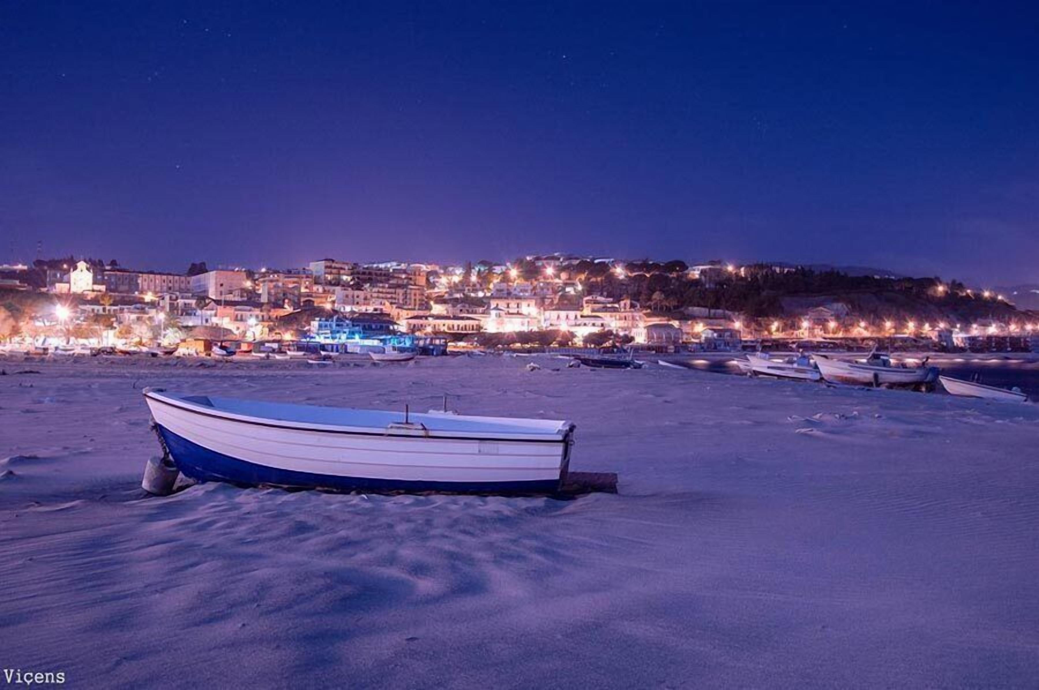 Escena costera con una hilera de barcos en una playa arenosa, un pueblo al fondo con edificios y luces, y un cielo nocturno despejado.