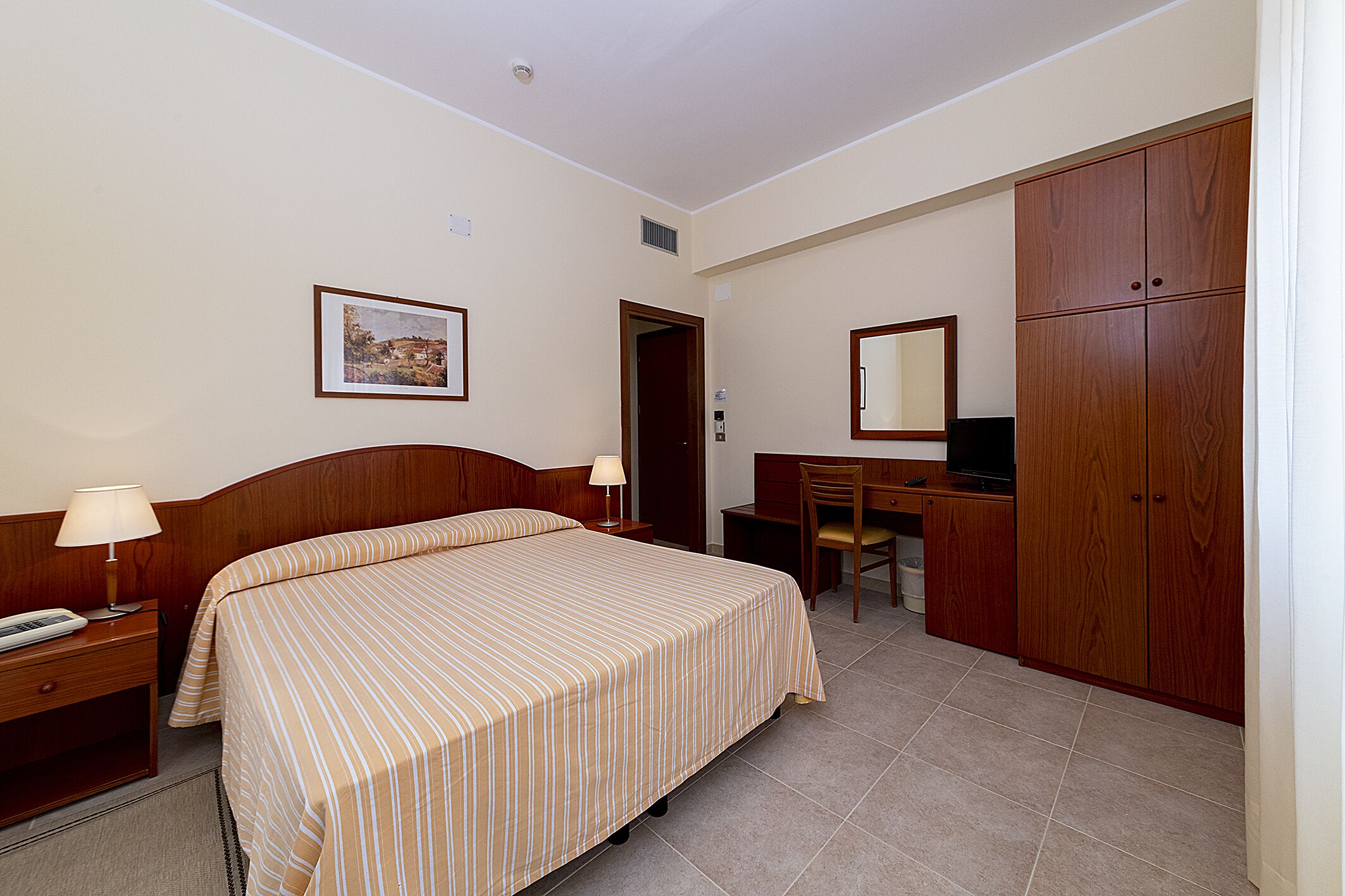 A hotel room with a bed, bedside table, lamp, desk, chair, and wardrobe.