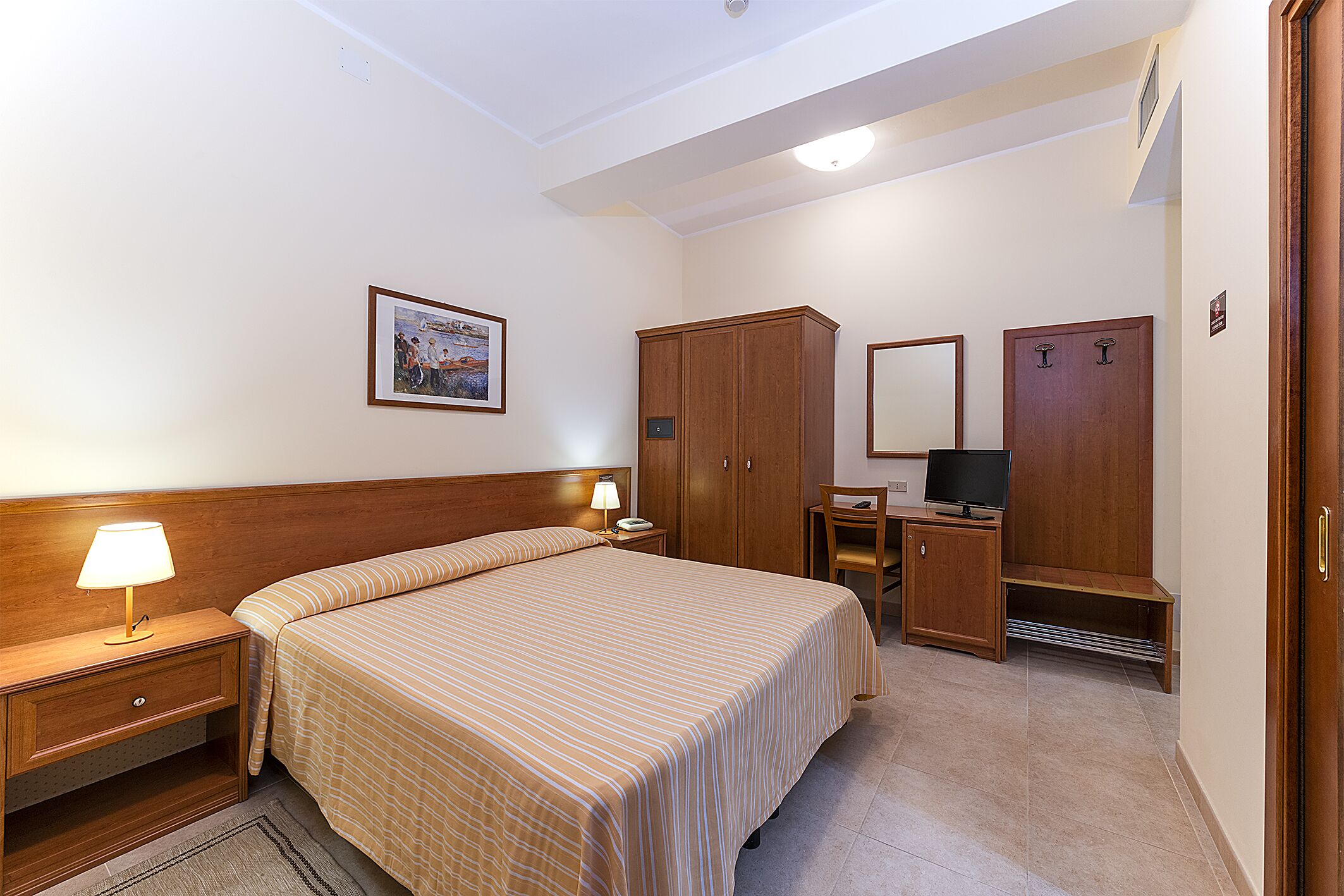 A hotel room with a bed, bedside table with a lamp, a desk with a chair, a wardrobe, and a television.