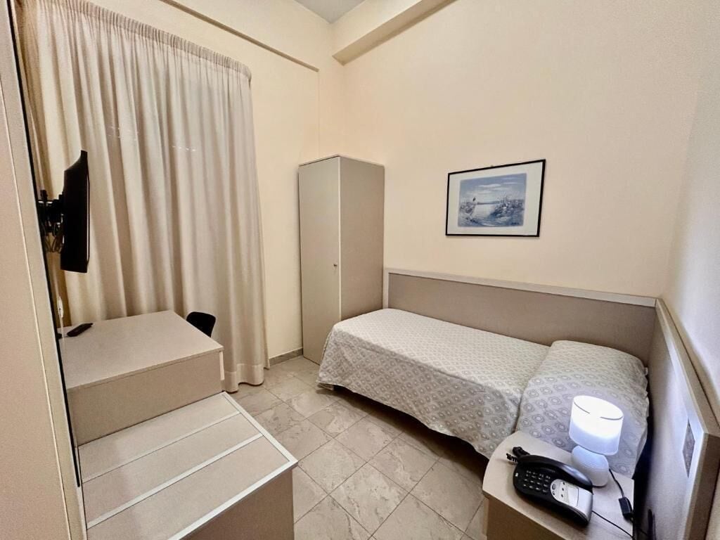 A compact hotel room with a bed, a desk, a chair, a wardrobe, a bedside table with a lamp, and a phone.