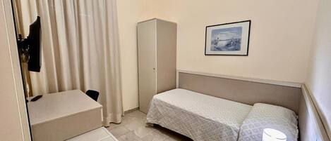 A compact hotel room with a bed, a desk, a chair, a wardrobe, a bedside table with a lamp, and a phone.