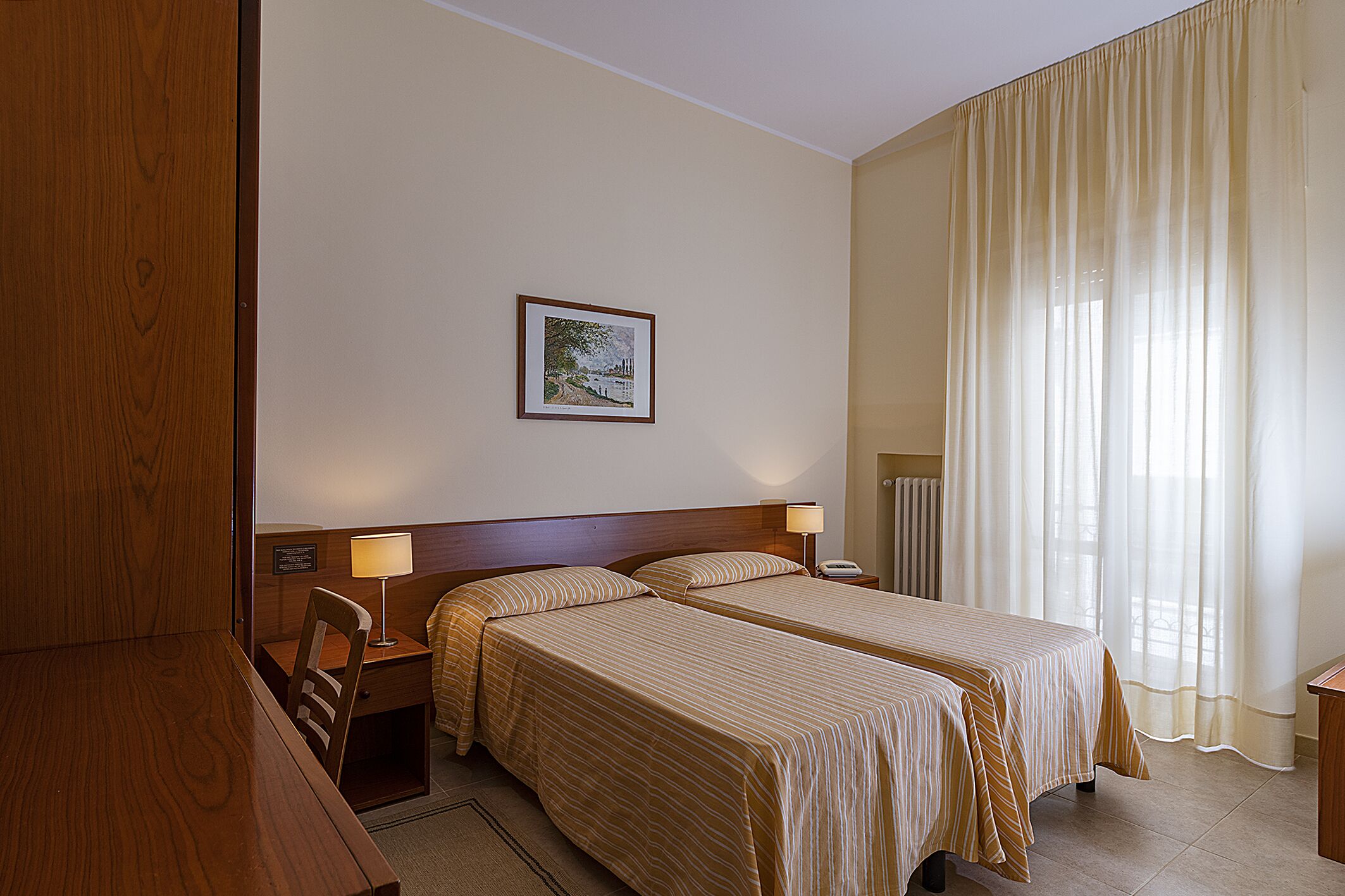 Une chambre d’hôtel avec deux lits, un bureau, une chaise, un tableau encadré au mur et une fenêtre avec des rideaux.